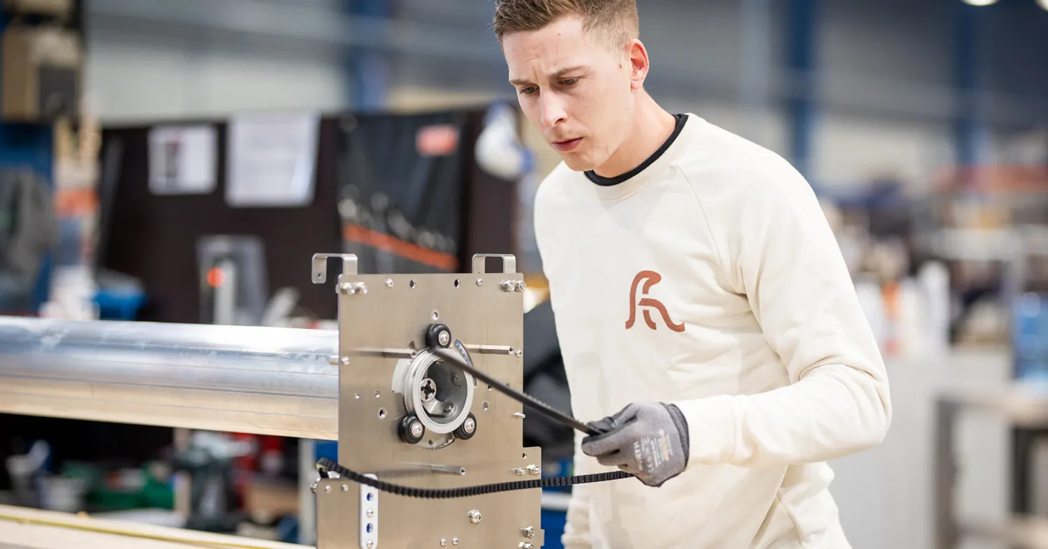 Un homme en pull blanc travaille sur une machine métallique dans un atelier industriel lumineux.