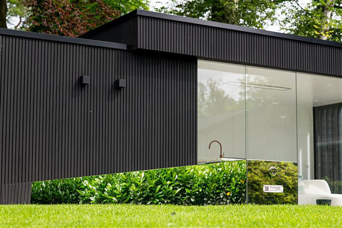 Une maison moderne en bois sombre avec de grandes fenêtres en verre, entourée de verdure et d'une pelouse verte.