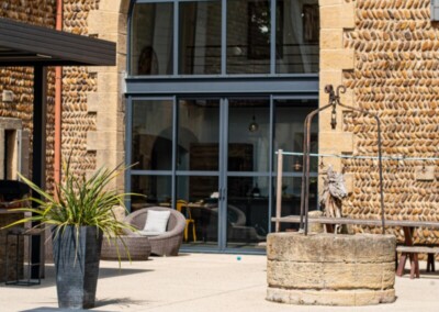 Façade en pierre avec grande fenêtre arquée, terrasse avec mobilier de jardin, plante en pot et piscine couverte par une structure grise.