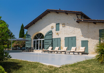 Perspective rapprochée sur la piscine recouverte par une Coverseal grise, devant une maison de campagne avec volets verts et terrasse ensoleillée.