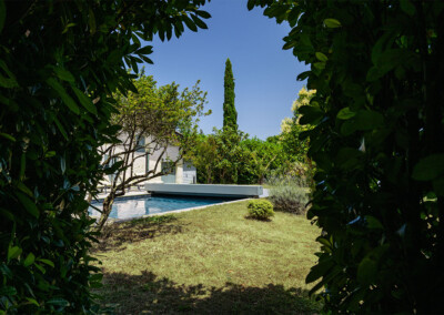 Vue depuis le jardin encadrée par des feuillages, laissant apparaître la piscine et sa couverture Coverseal grise au cœur d’un écrin de verdure.