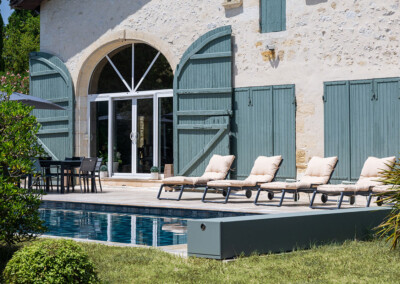 Zoom sur la piscine protégée par une Coverseal grise, au pied d’une maison en pierre avec grande baie vitrée et volets verts.