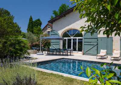 Vue sur la piscine ouverte, au pied d’une maison en pierre et mobilier d’extérieur sous parasols gris.